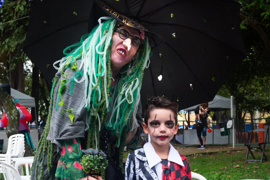 Bruxa de cabelos verdes e guarda-chuva preto posando com um menino fantasiado de palhaço no Festival Halloween Moema
