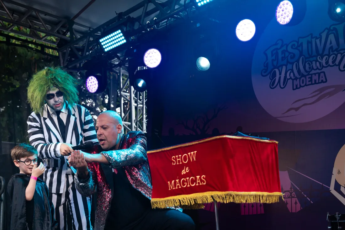 Mágico de jaqueta de paetês apresentando um show de mágicas no palco do Festival Halloween Moema, com uma criança assistindo